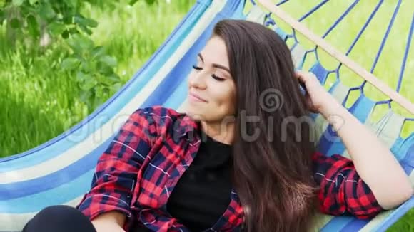 一个美丽的快乐女孩的特写肖像躺在花园的吊床上微笑和放松视频的预览图