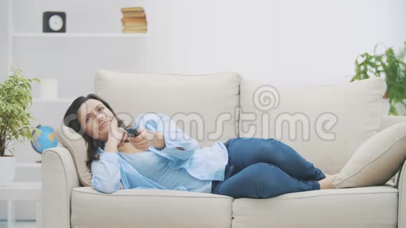 年轻放松的女人正在用Zapper切换频道她正在看电视躺在家里的沙发上复制空间4K慢慢视频的预览图