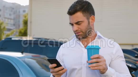 办公大楼附近的停车场上有一个英俊的男人使用智能手机和饮用美味的咖啡看上去很满意视频的预览图