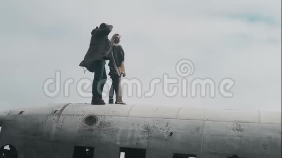 一对年轻的旅行夫妇站在冰岛坠毁的DC3飞机上在大风天一起举手视频的预览图