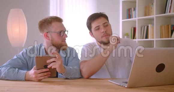 两位成年白种人商人使用笔记本电脑和平板电脑在室内进行讨论的特写照片视频的预览图