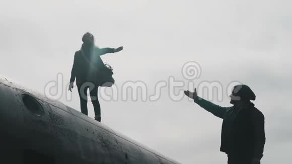 年轻女子站在冰岛坠毁的DC3飞机的顶部伸手向站在附近的男人伸出手视频的预览图