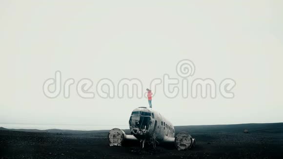 穿着粉色夹克的年轻女旅行者站在冰岛坠毁的DC3飞机上环顾着黑色的海滩视频的预览图