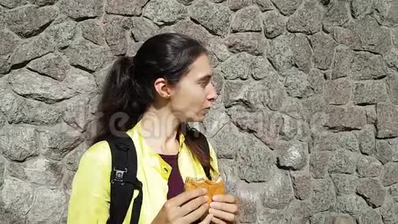 布鲁内特女孩旅行者穿着夹克和背包在街上吃着一个面包旅途中的快餐微笑和外表视频的预览图