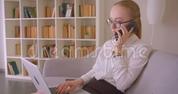 特写侧景照片照片中年轻漂亮的白种人金发女商人戴着眼镜使用笔记本电脑和手机视频的预览图