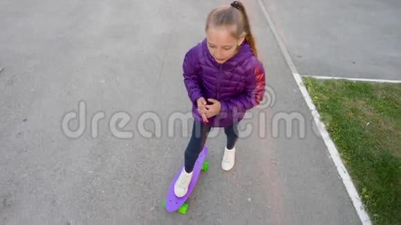 穿着夹克的快乐女孩在公园的街道上的紫色滑板上滚动视频的预览图