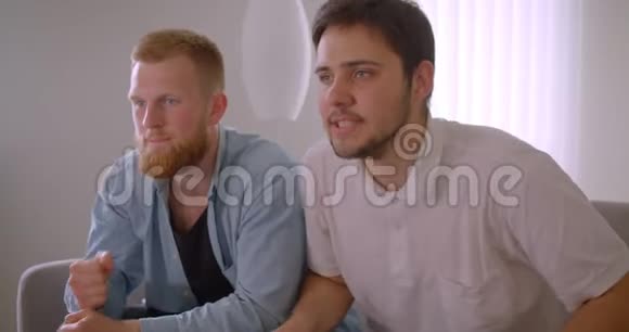 两位英俊的白种人成年男子在电视上观看体育节目并为他们的足球队坐着欢呼视频的预览图