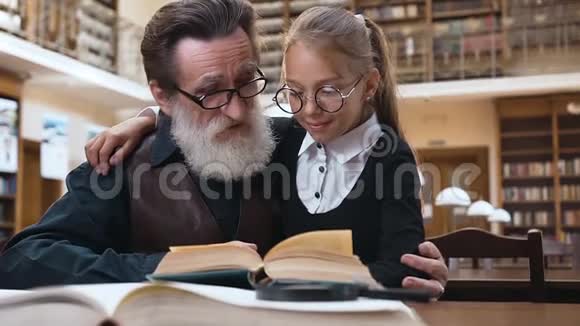 可爱的一对孙女和她的老胡子祖父的漂亮肖像坐在图书馆和视频的预览图