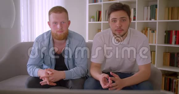 两位英俊的白种人成年男子在电视上看体育节目坐在室内沙发上放松视频的预览图