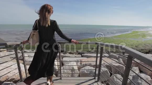 穿着黑色裙子的女人被风吹动看着地平线上的大海视频的预览图