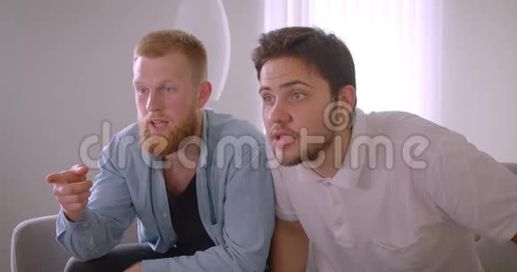 两位英俊的白种人成年男子在电视上观看体育节目高兴地坐在沙发上庆祝视频的预览图