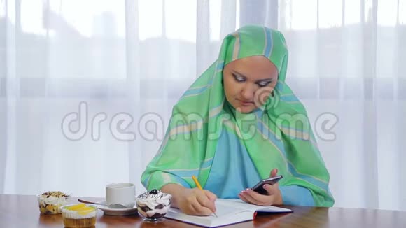 一个年轻的穆斯林妇女在一家咖啡馆里戴着浅绿色的围巾用电话和日记远程工作视频的预览图