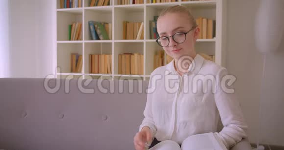 年轻漂亮的白种人金发女学生戴着眼镜看着一本书微笑着看着摄像机的特写照片视频的预览图