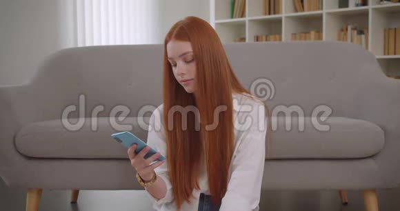 年轻可爱的红发女郎用手机对着坐在地板上的相机微笑的特写照片视频的预览图
