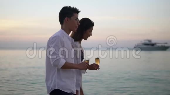 年轻快乐的一对夫妇手里拿着一杯香槟在海滩上散步美丽温暖的夜晚在海边视频的预览图
