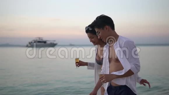 美丽的年轻夫妇在日落时散步由海洋比基尼女孩和英俊的男孩拿着酒杯欣赏着视频的预览图