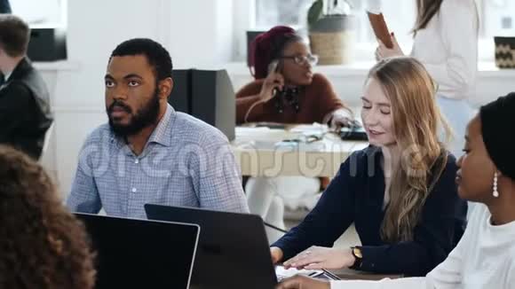 积极的年轻金发女人和英俊的非洲男人坐在办公室桌上在现代的灯光合作视频的预览图