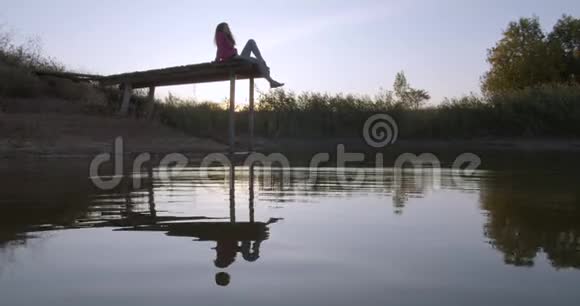 女人在湖边放松坐在一个木制码头的边缘在摆动腿视频的预览图