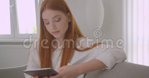 年轻漂亮红发女郎浏览平板电脑看着相机兴高采烈地坐在上面的特写照片视频的预览图