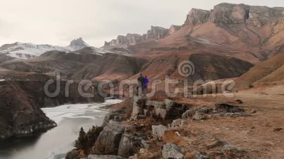 一对年轻夫妇站在靠近山湖的悬崖边上以史诗般的岩石为背景的鸟瞰图视频的预览图