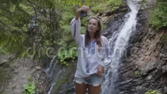 可爱的年轻女子穿着刺绣衬衫自拍站在瀑布前与野性的联系休闲视频的预览图