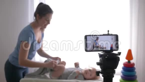工作博客作者年轻妈妈vlogger在智能手机上录制教育视频时给蹒跚学步的男孩换衣服视频的预览图