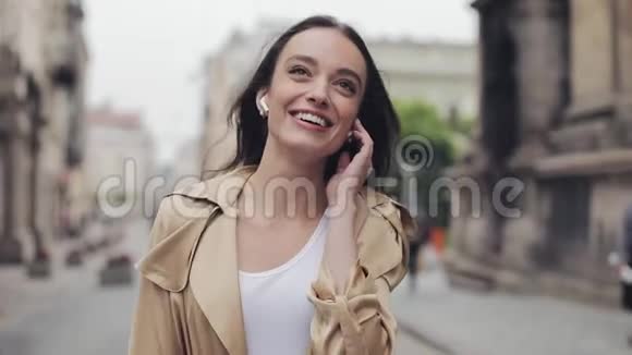 美丽快乐的年轻女孩用无线耳机说话的肖像微笑着在街上轻松地散步视频的预览图