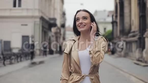 美丽快乐的年轻女孩用无线耳机说话的肖像微笑着在街上轻松地散步视频的预览图