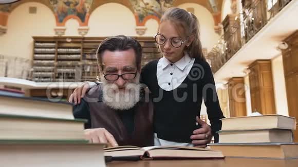 聪明的老胡子爷爷戴着眼镜看书和他可爱的小孙女在图书馆的桌子上视频的预览图