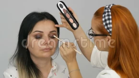美丽的红发化妆师在一家美容院为一位年轻女子化妆调色盘为米色眼影视频的预览图
