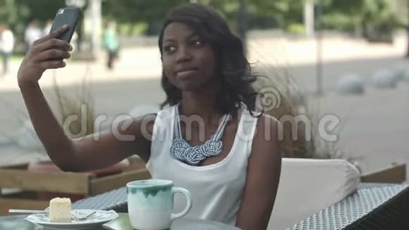 迷人的非洲年轻女子微笑着独自坐在户外咖啡馆里用智能手机自拍视频的预览图
