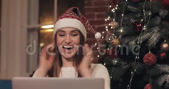 年轻漂亮的女人戴着圣诞帽坐在沙发上在家里背景下用她的笔记本电脑开心地笑着视频的预览图