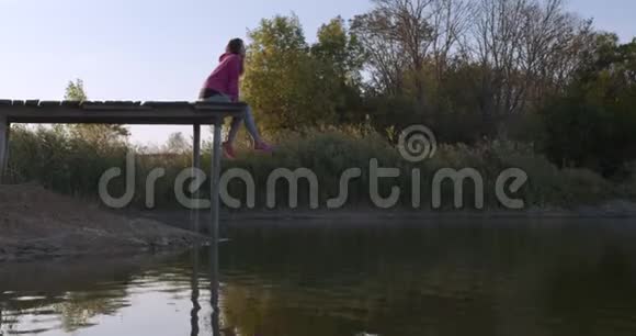 女人在湖边放松坐在一个木制码头的边缘在摆动腿视频的预览图