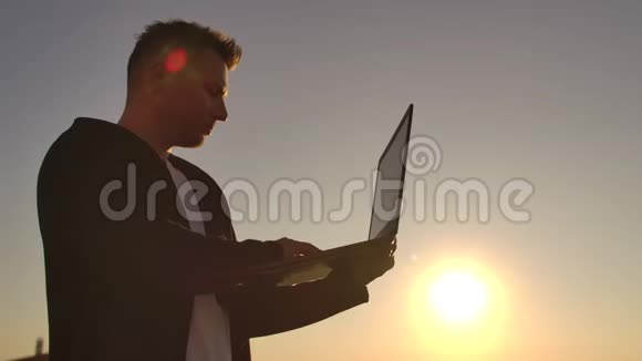 中期计划一名男性自由职业者在日落时分在笔记本电脑键盘上打字俯瞰城市同时站在城市的屋顶上视频的预览图