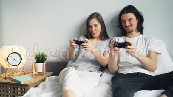 可爱的年轻夫妇男朋友和女朋友在家里的床上玩电子游戏视频的预览图