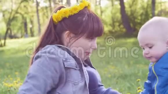 年轻女子头上戴着一个蒲公英花圈在草地上和穿着蓝色夹克的小儿子一起玩视频的预览图