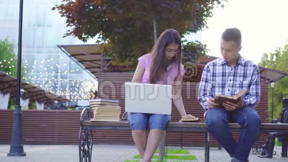年轻的亚洲学生男人拿着书女人拿着笔记本电脑坐在长凳上视频的预览图