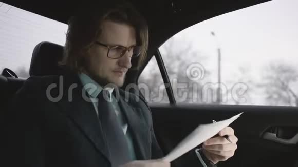 漂亮的男人棕色头发穿着开心果衬衫灰色西装戴着眼镜坐在车里看文件视频的预览图