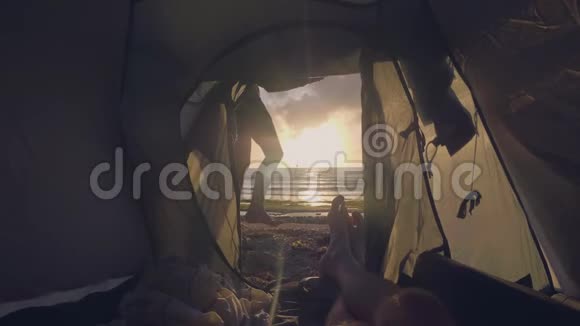 从旅游帐篷的轮廓看年轻的女孩在金色的日落景观在海上和男性的脚躺在里面观点看法视频的预览图