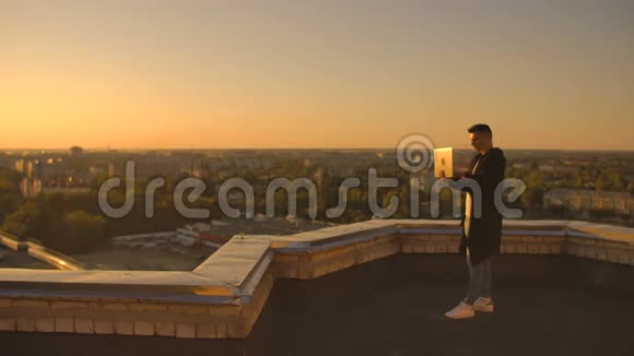 中期计划一名男性自由职业者在日落时分在笔记本电脑键盘上打字俯瞰城市同时站在城市的屋顶上视频的预览图