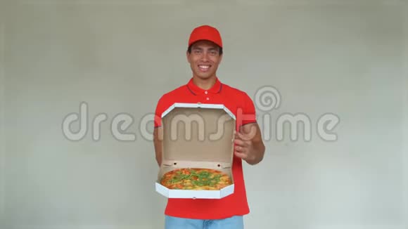 身穿红色制服的英俊年轻送货员正拿着一个披萨看着相机微笑着灰色背景视频的预览图