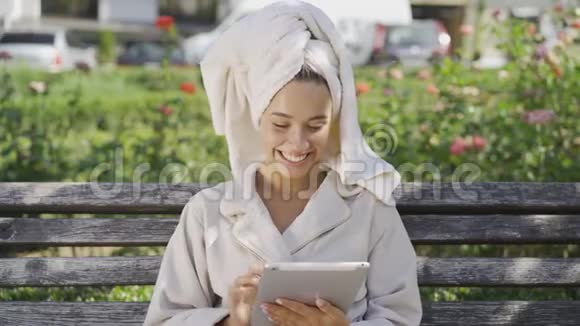 一幅身着浴袍头戴毛巾坐在公园长椅上查看资料的微笑年轻女子的画像视频的预览图