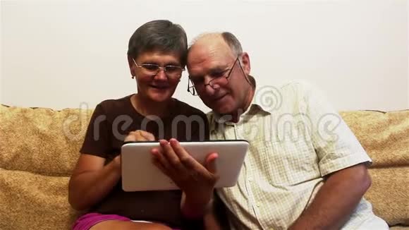 一对老夫妇正坐在家里的沙发上在平板电脑上看照片微笑着积极地交谈视频的预览图