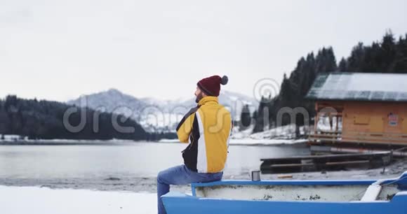 在一个白雪皑皑的湖泊和森林旁边一位魅力非凡的年轻游客欣赏他坐在一艘蓝色小船旁边的景色视频的预览图