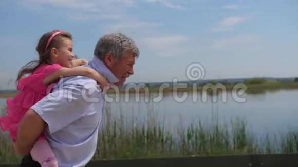 一个小女孩和她的祖父坐在她的背上他们沿着湖跑4K慢慢视频的预览图