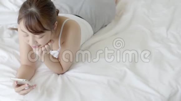 美丽的年轻亚洲女人在卧室里享受着床上的乐趣看着摸着智能手机阅读社交网络视频的预览图