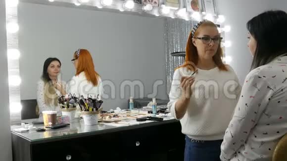 美丽的红发化妆师在一家美容院为一位年轻女子化妆调色盘为米色眼影视频的预览图