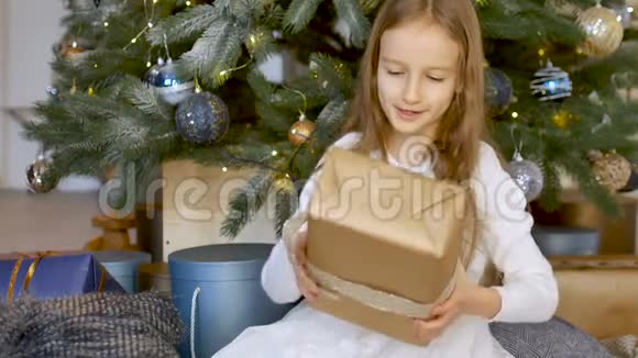 可爱的金发女孩收到了一个礼品盒里面包着工艺纸坐在圣诞树附近快乐的童年和圣诞节视频的预览图