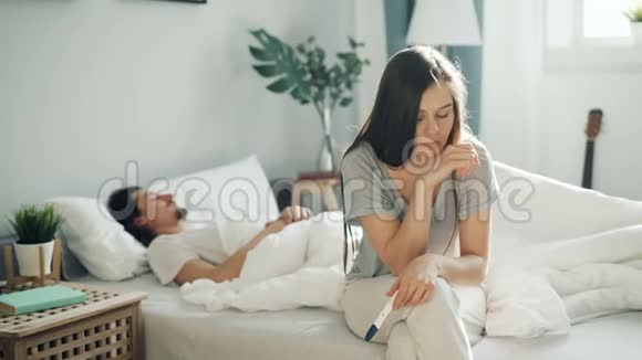 紧张的女孩坐在床上拿着怀孕测试而男人睡在卧室里视频的预览图