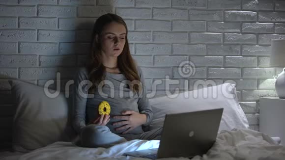 年轻孕妇在笔记本电脑前哭泣吃甜甜圈以克服压力视频的预览图